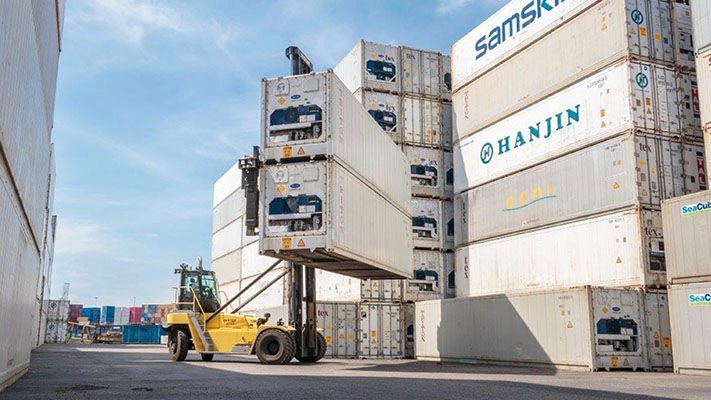 Top-loading container handler at a dock removing two very large shipping containers from a stack of containers.